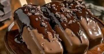 homemade chocolate covered ice cream bars with glossy chocolate shell and chocolate shavings on top placed on parchment paper with wooden sticks visible.