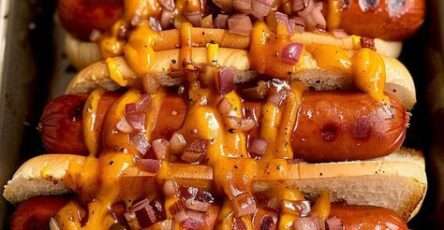 Grilled hot dogs topped with melted cheddar cheese sauce and diced red onions served in toasted brioche buns on a wooden serving board.