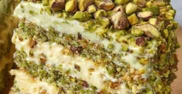 Two-layer pistachio cake filled and frosted with mascarpone cream and topped with chopped pistachios on a white cake stand.
