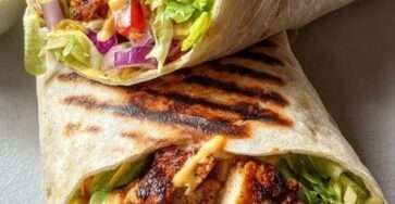 Grilled chicken wraps filled with lettuce, tomatoes, red onion and creamy garlic sauce wrapped in warm flour tortillas on a wooden plate.
