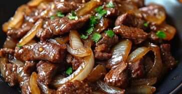 Pan-fried beef strips with caramelized onions in a skillet, coated in a glossy soy-based sauce and garnished with fresh parsley, served hot.
