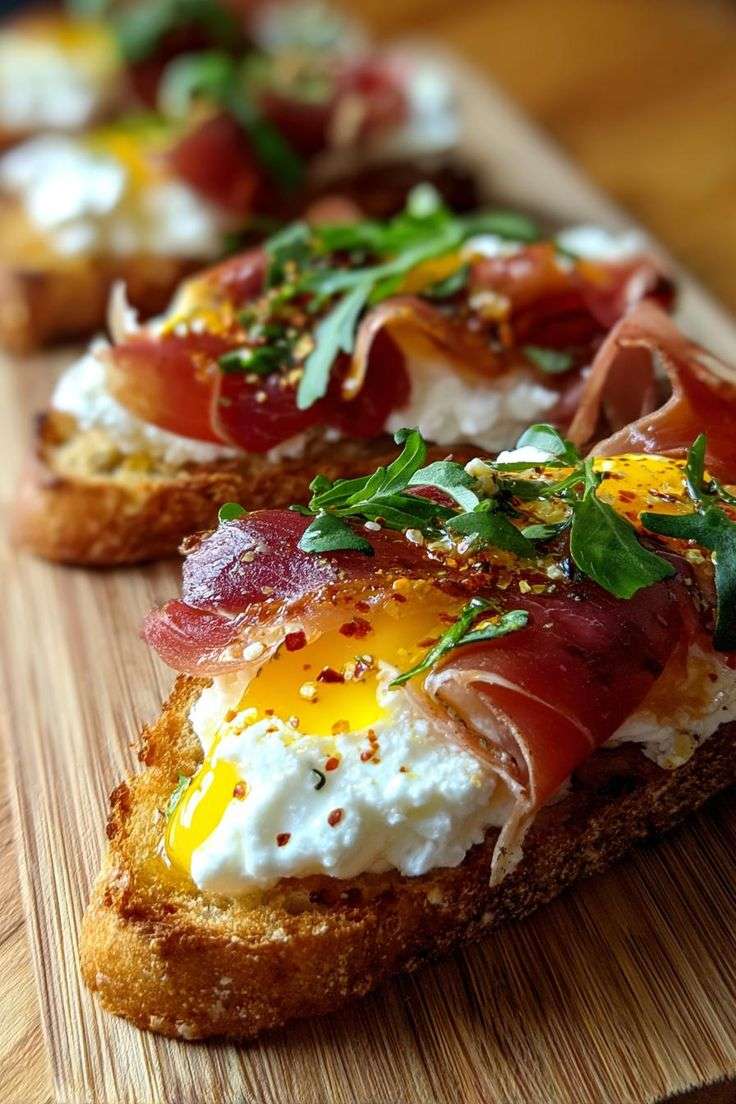 Crispy toasted crostini topped with creamy burrata, delicate prosciutto, poached eggs, fresh arugula, red pepper flakes, olive oil, and balsamic glaze on a rustic serving platter.