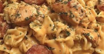 A bowl of crockpot cheddar ranch chicken and sausage pasta with creamy sauce, melted cheese, sliced sausage, and tender chicken pieces garnished with parsley.