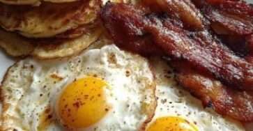 A classic American breakfast plate with a stack of fluffy pancakes topped with butter and maple syrup, crispy bacon strips, and sunny-side-up eggs with runny yolks.