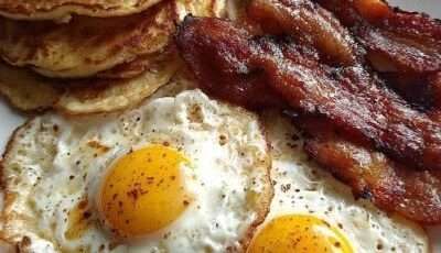 A classic American breakfast plate with a stack of fluffy pancakes topped with butter and maple syrup, crispy bacon strips, and sunny-side-up eggs with runny yolks.