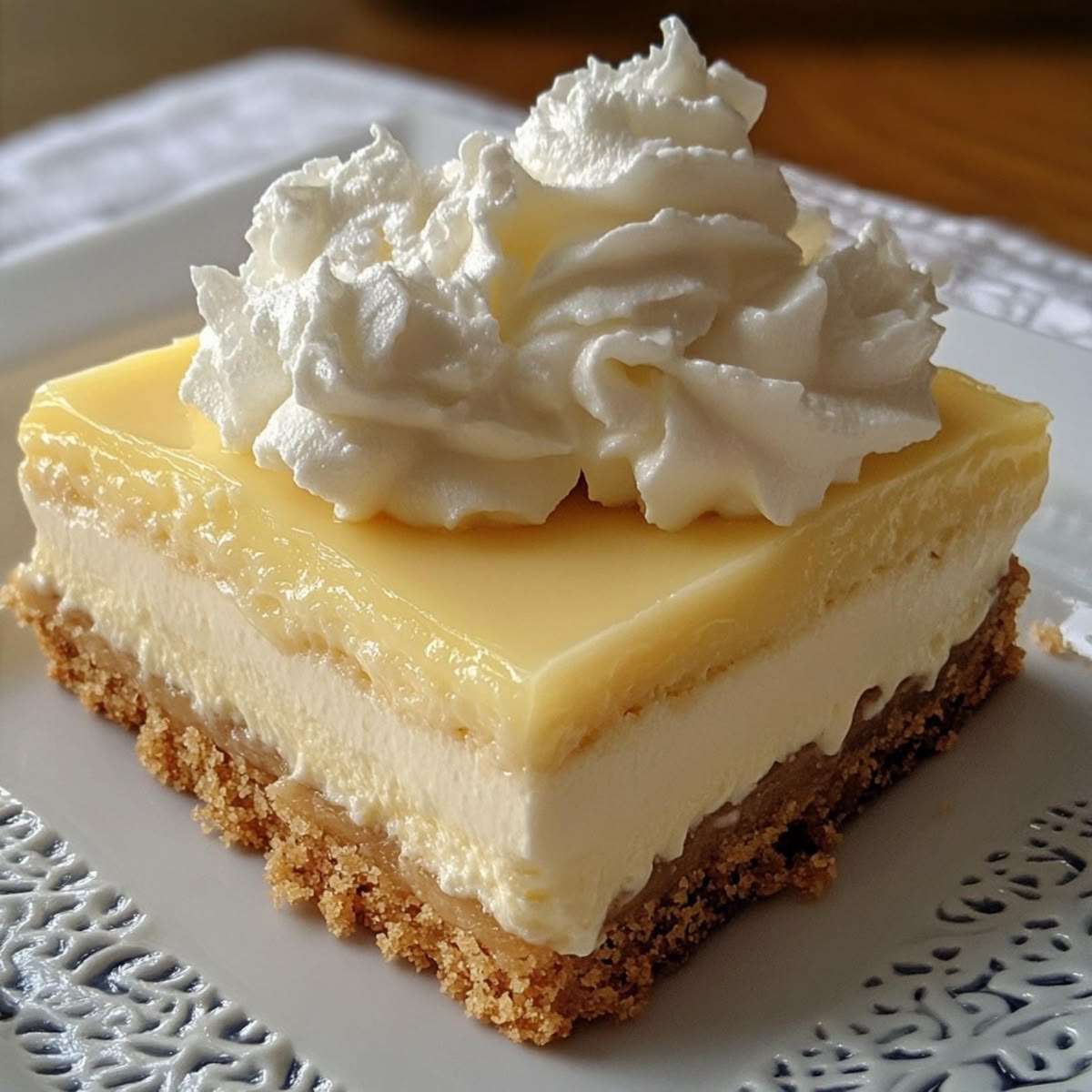 No-bake lemon cheesecake bars with a buttery biscuit crust, creamy cheesecake layer, glossy lemon curd topping, and whipped cream decoration on top.