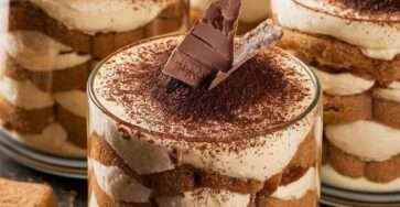 No-bake tiramisu cups in glass jars with layers of coffee-soaked ladyfingers, creamy mascarpone filling, cocoa powder dusting, and chocolate shavings on top.