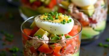 Layered tuna avocado verrines in glass cups with creamy mashed avocado, seasoned tuna, diced tomatoes, and half boiled egg on top, garnished with chives.