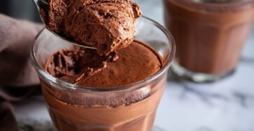 Traditional chocolate mousse served in glass cups with a smooth, airy texture, topped with chocolate shavings and a slightly glossy surface, placed on a dark elegant background.