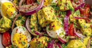 A clear glass bowl filled with a colorful avocado and egg salad, showcasing hard-boiled egg quarters, diced green avocado, red onion slices, and cherry tomatoes.