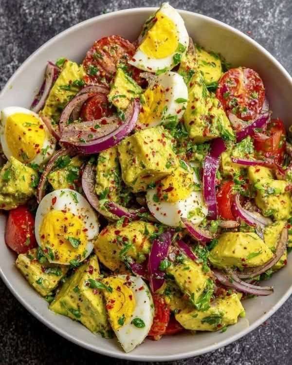 A clear glass bowl filled with a colorful avocado and egg salad, showcasing hard-boiled egg quarters, diced green avocado, red onion slices, and cherry tomatoes.
