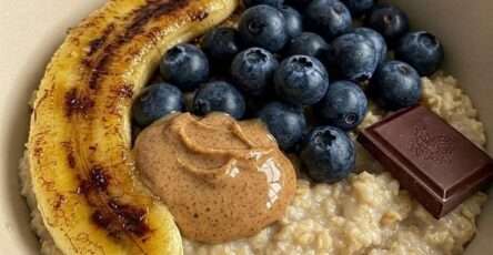 Creamy oatmeal bowl topped with caramelized banana halves, fresh blueberries, peanut butter, dark chocolate, and a drizzle of honey.