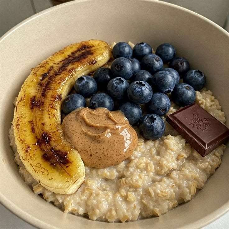 Creamy oatmeal bowl topped with caramelized banana halves, fresh blueberries, peanut butter, dark chocolate, and a drizzle of honey.
