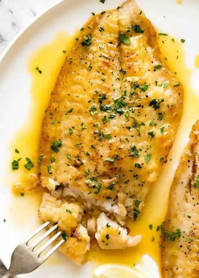 Lemon butter fish fillets served with glossy garlic lemon butter sauce, fresh parsley, lemon wedges, and a side of vegetables.