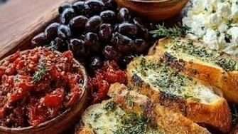 Mediterranean appetizer board with crispy garlic toast, black olives, green olives, feta cheese, sun-dried tomatoes, roasted pepper spread, and parsley.
