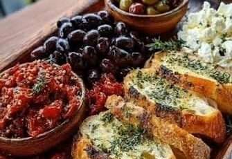 Mediterranean appetizer board with crispy garlic toast, black olives, green olives, feta cheese, sun-dried tomatoes, roasted pepper spread, and parsley.