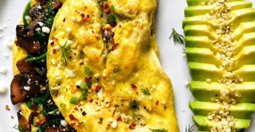 Mushroom spinach omelette served with sliced avocado, crumbled feta cheese, fresh herbs, sesame seeds, black pepper, and a small squeeze of lemon.