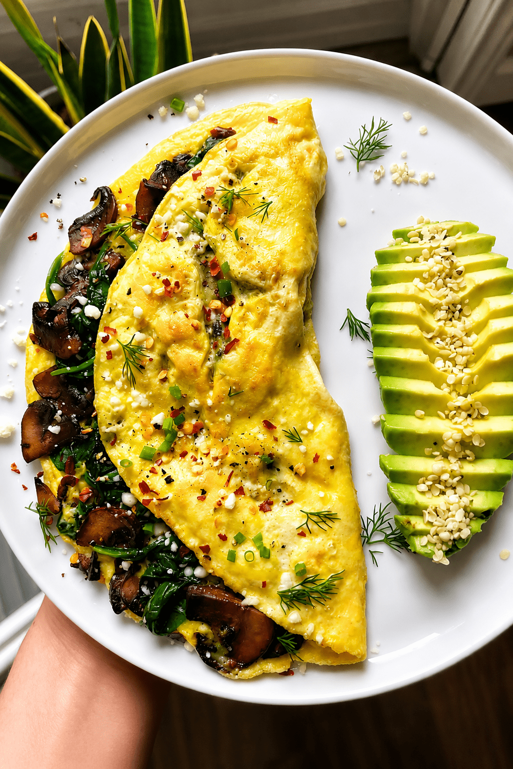 Mushroom spinach omelette served with sliced avocado, crumbled feta cheese, fresh herbs, sesame seeds, black pepper, and a small squeeze of lemon.
