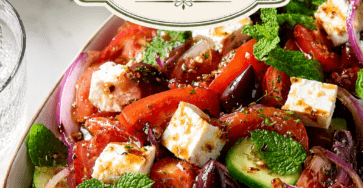 Fresh Greek feta cucumber tomato salad served in a bowl with tomato wedges, cucumber slices, red onion, feta cubes, olives, mint, parsley, and oregano dressing.