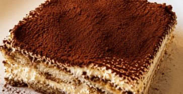 A classic tiramisu slice served on a plate with creamy mascarpone layers, coffee-soaked ladyfingers, a thick dusting of cocoa powder, and dark chocolate shavings.