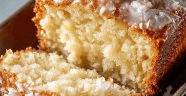 A moist coconut loaf cake sliced on a plate with creamy white glaze, shredded coconut topping, soft tender crumb, whipped cream, and fresh mint garnish.