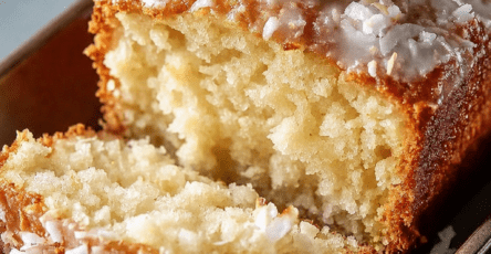 A moist coconut loaf cake sliced on a plate with creamy white glaze, shredded coconut topping, soft tender crumb, whipped cream, and fresh mint garnish.