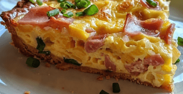 A golden ham and cheese quiche sliced on a plate with creamy egg filling, diced ham, melted cheese, fresh chives, flaky crust, and black pepper on top.