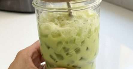 A tall mason jar filled with creamy iced matcha latte made with oat milk and nugget ice. The vibrant green matcha layer swirls beautifully through the milk, creating a marbled effect. The drink is served with a metal straw and lid, making it a refreshing and aesthetic homemade beverage.