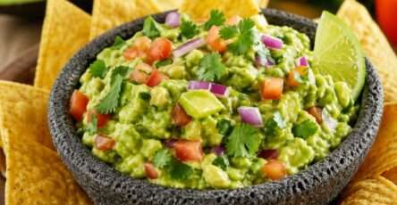 Classic homemade guacamole served in a bowl with ripe avocado, diced tomato, red onion, cilantro, lime wedges, and tortilla chips.