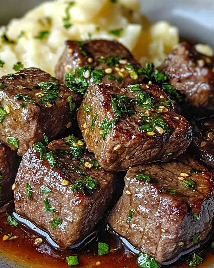 Garlic butter steak bites served over creamy mashed potatoes with garlic butter sauce, fresh parsley, sesame seeds, and black pepper.