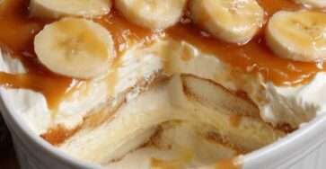 A rectangular baking dish showing layers of soft cookies, fluffy white cream, and sliced bananas, all topped with a generous drizzle of glossy golden caramel sauce.