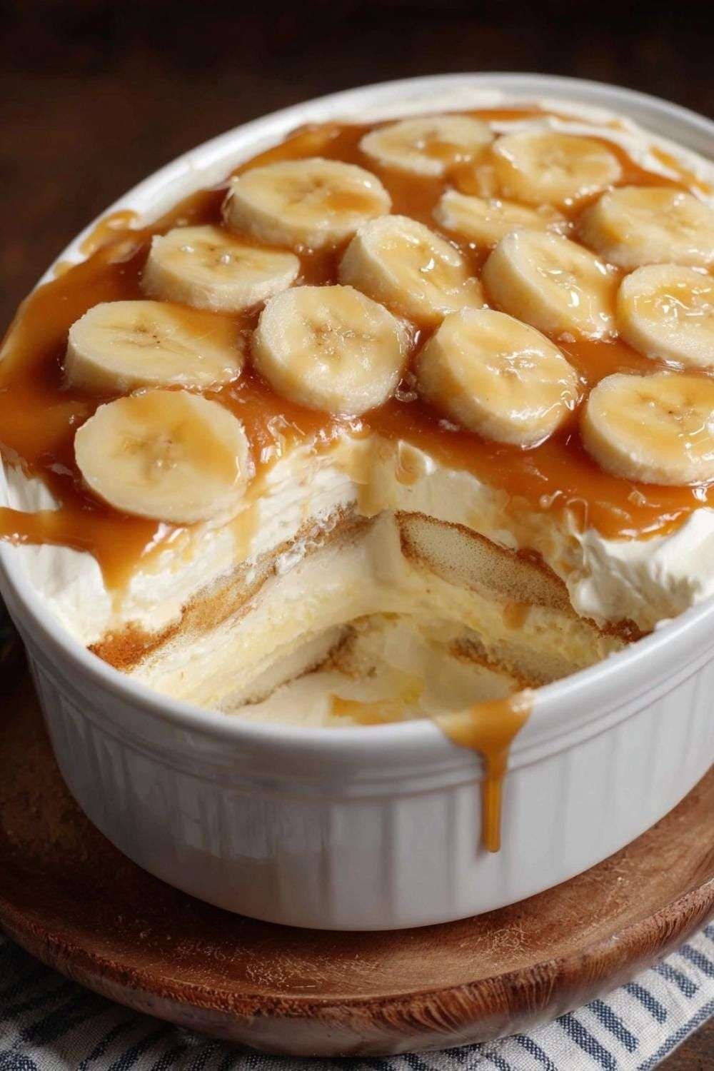 A rectangular baking dish showing layers of soft cookies, fluffy white cream, and sliced bananas, all topped with a generous drizzle of glossy golden caramel sauce.