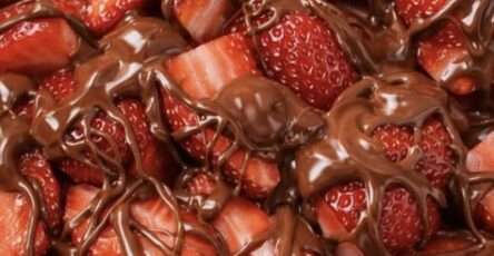 A white ceramic bowl filled with fresh red strawberry halves heavily drizzled with shiny melted dark chocolate against a clean white background.