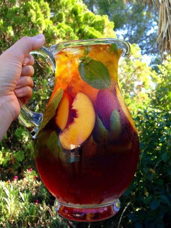 A large glass pitcher of peach iced tea with sliced peaches, fresh mint leaves, lemon juice, and ice cubes served cold in clear glasses.