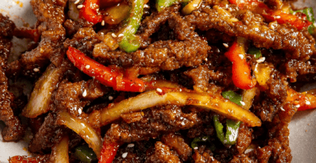Crispy chilli beef stir-fry served with sliced bell peppers, onion, sesame seeds, and a glossy sweet spicy sauce over rice.