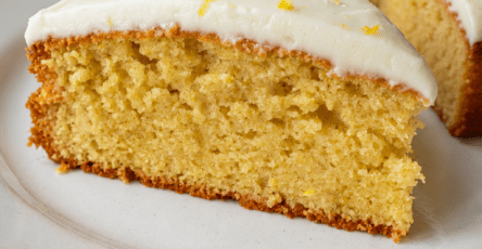 A soft lemon cake topped with smooth cream cheese frosting, fresh lemon zest and a moist golden crumb served on a cake plate.