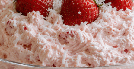 A glass bowl of sugar-free strawberry coconut cheesecake fluff topped with whole strawberries and shredded coconut, with a creamy pink texture.