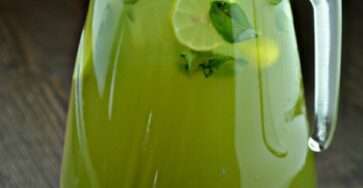 A large pitcher of fresh mint lemonade with lemon slices, lime slices, fresh mint leaves, cold water, and ice cubes served in clear glasses.