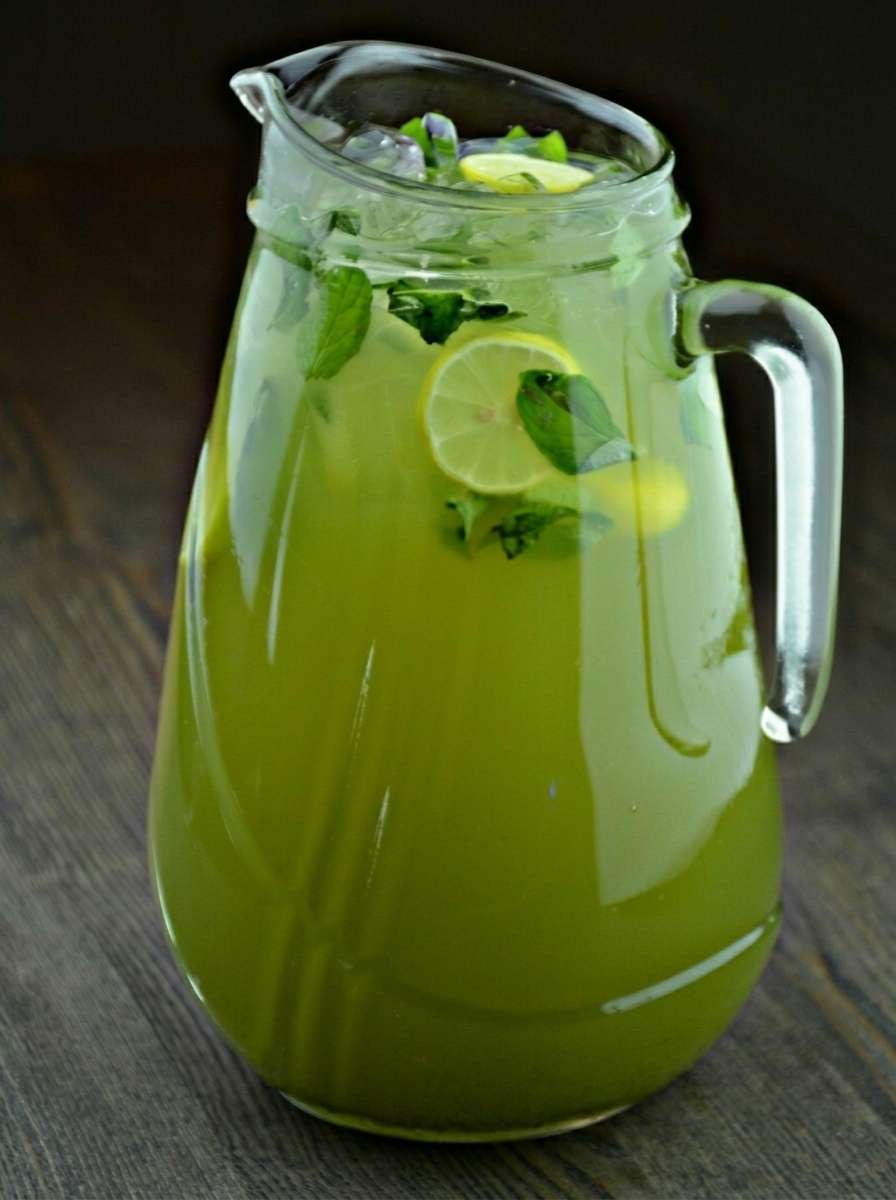 A large pitcher of fresh mint lemonade with lemon slices, lime slices, fresh mint leaves, cold water, and ice cubes served in clear glasses.