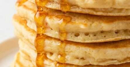 A stack of homemade fluffy pancakes on a plate topped with a pat of butter and golden pancake syrup dripping over the sides.