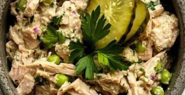 Creamy tuna salad served in a bowl with green peas, sliced pickles, red onion, parsley, and a smooth lemon mayonnaise dressing.