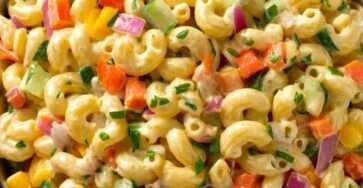 A colorful pasta salad with macaroni, diced carrots, red and yellow bell peppers, cucumber, red onion, creamy dressing, and fresh parsley in a large bowl.
