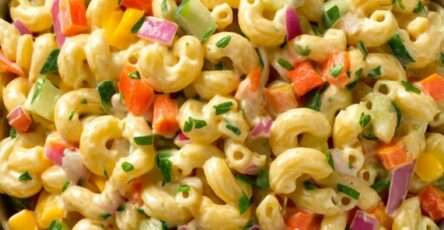 A colorful pasta salad with macaroni, diced carrots, red and yellow bell peppers, cucumber, red onion, creamy dressing, and fresh parsley in a large bowl.