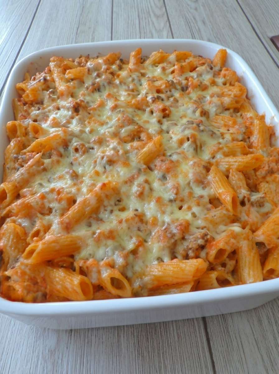A golden baked pasta gratin with ground meat, tomato sauce, melted cheese, penne pasta, and fresh parsley in a rustic oven dish.