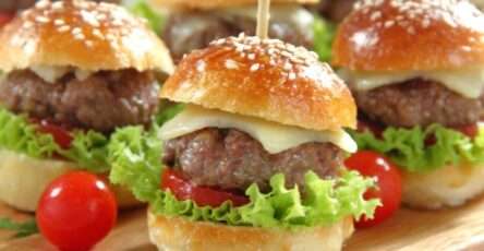 Gourmet mini burgers with juicy beef patties, melted cheese, lettuce, cherry tomatoes, toasted buns, and wooden skewers on a serving plate.