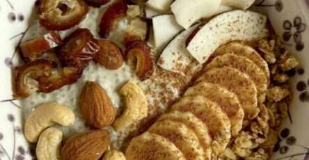 A creamy yogurt chia breakfast bowl topped with banana slices, apple or pear slices, chopped dates, granola, almonds, cashews, coconut slices, and cinnamon.