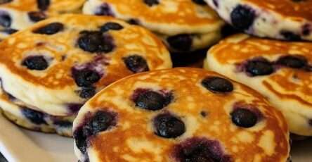 A stack of fluffy blueberry pancakes served warm with juicy blueberries, golden edges, soft thick texture, butter, and maple syrup on top.