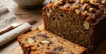 Sliced homemade date and walnut loaf topped with crunchy walnuts and a glossy honey glaze, served on a wooden board beside a warm cup of tea.