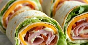 Ham and cheese tortilla roll-ups sliced into pinwheels with creamy cheese spread, cooked ham, cheddar, lettuce, and tomatoes on a serving plate.