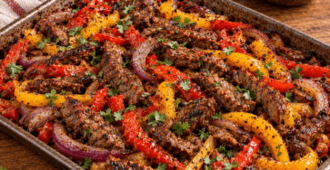 Sheet pan fajita-style beef with roasted red and yellow peppers, sliced onions, warm tortillas, and creamy lemon sauce served on the side.